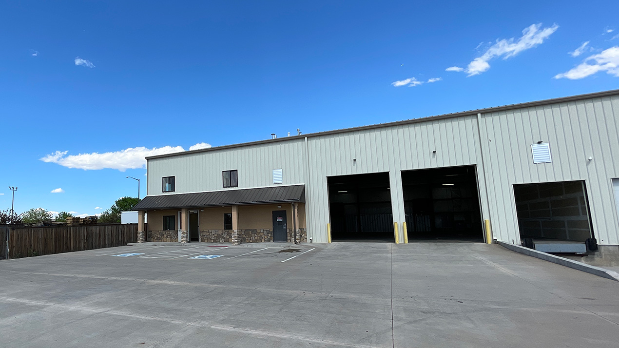 Industrial warehouse with two large loading bays and a small office on the left; blue sky overhead.