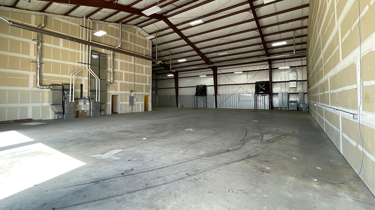 Empty industrial warehouse interior with beige drywall walls, exposed steel roof trusses, and large HVAC ductwork along the left wall.
