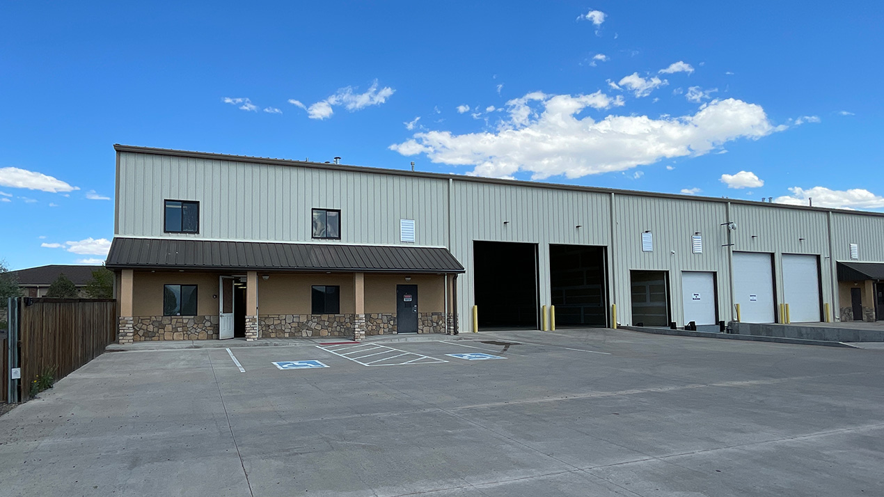 Front of a large warehouse: beige office section with stone trim, multiple loading bays and a wide concrete parking lot under a blue sky.