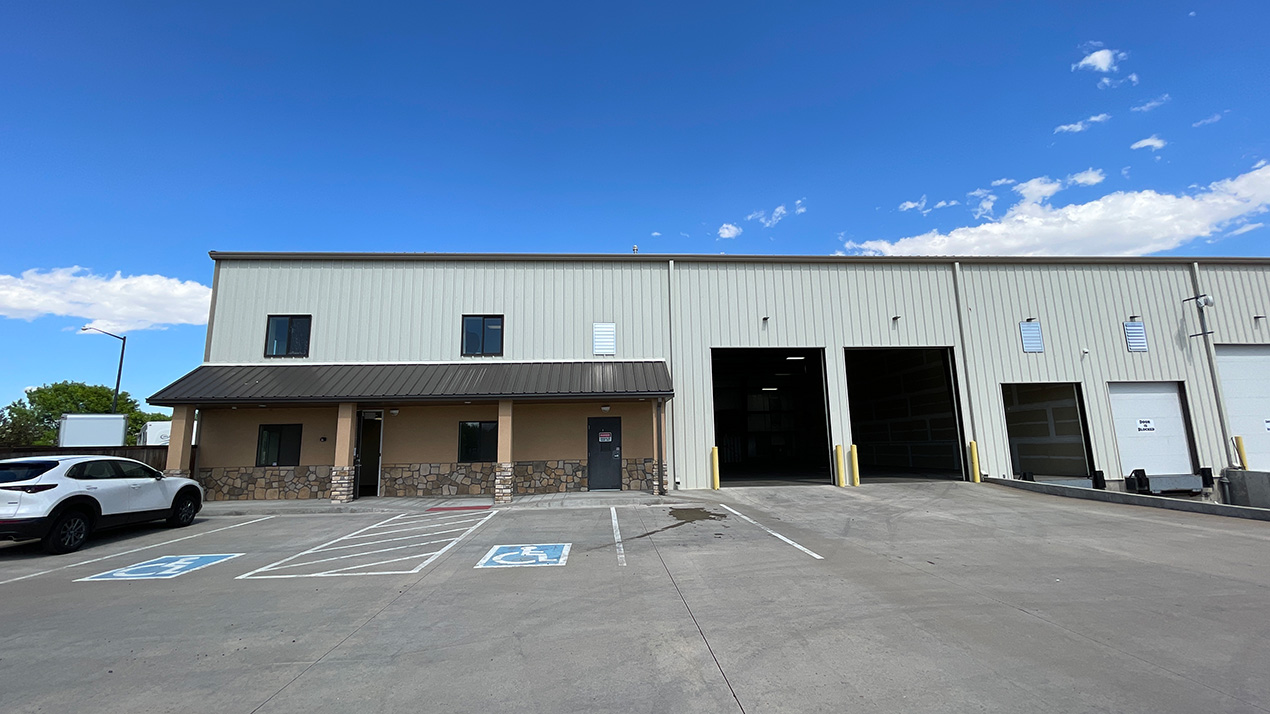 Industrial warehouse building with loading bays and a small storefront along stone facade, visible parking lot in foreground.