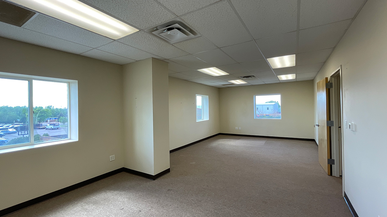 Empty office space with beige walls, carpet, and several ceiling lights under a drop ceiling, with windows on the far wall and a door to the right.