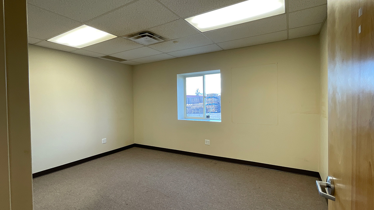 Empty small office with beige walls, brown baseboard, a single window, and a wooden door partly open.