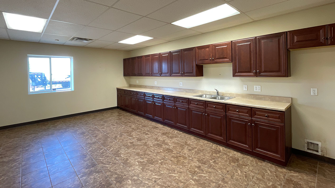 Long row of dark wood cabinets with beige countertops and a double sink along a wall in an empty kitchen room.
