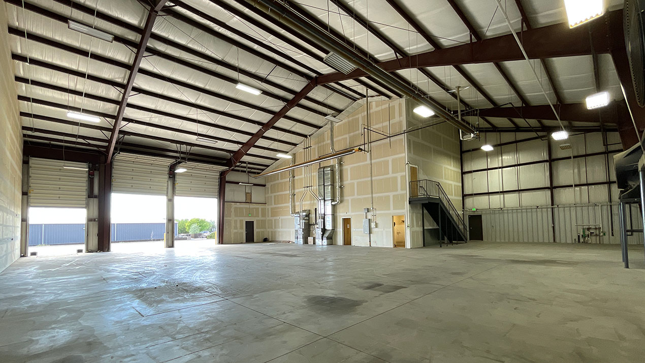 Empty industrial warehouse interior with tall ceilings, exposed steel beams, and large open bays; daylight through open loading doors to the outside.