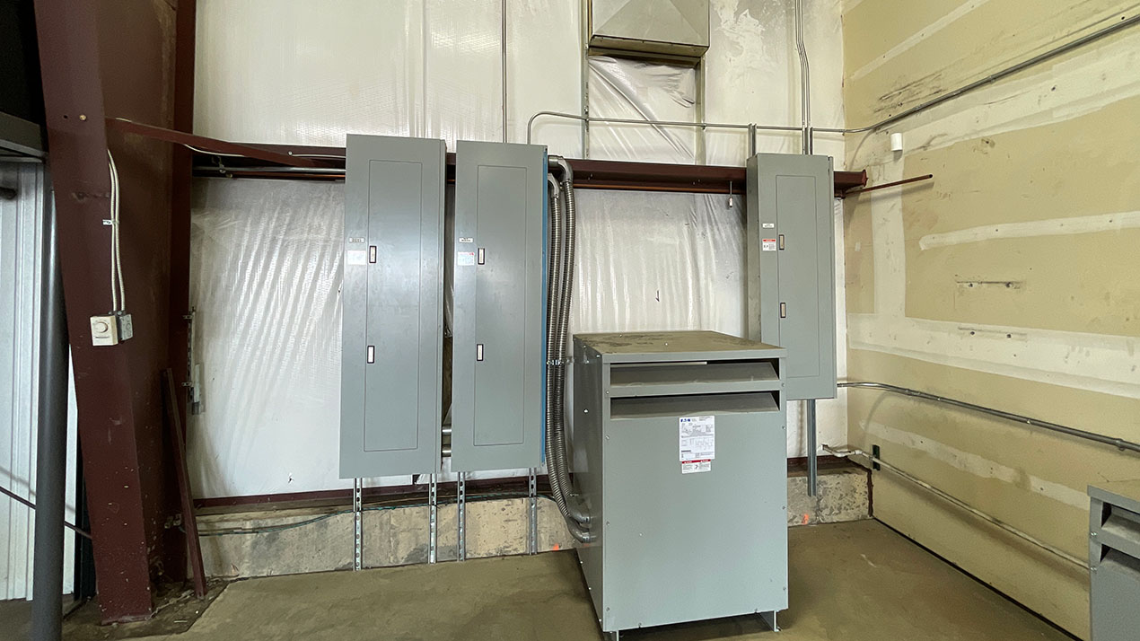 Industrial electrical room with four gray panels on the wall and a large gray cabinet in front, with conduits and wiring along the baseboard.